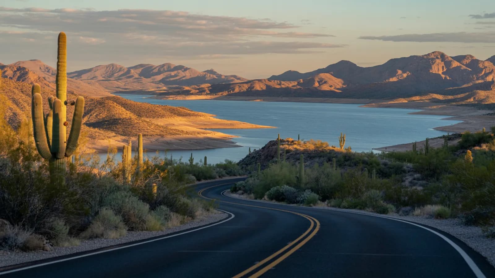 Arizona desert parkway winding toward a distant reservoir lake and rugged hills at golden hour